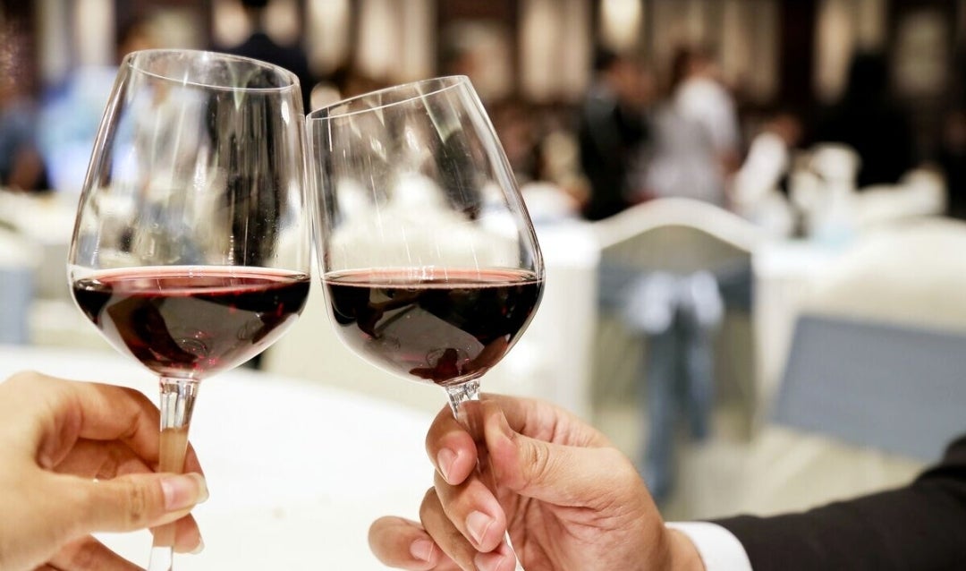 Two people holding glasses of red wine with a party in the background.