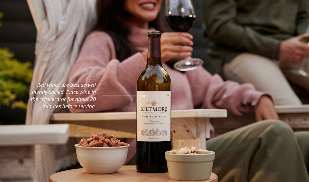 A bottle of red wine on a table with people socializing in the background. On the image is a tip recommending serving red wine slightly chilled.